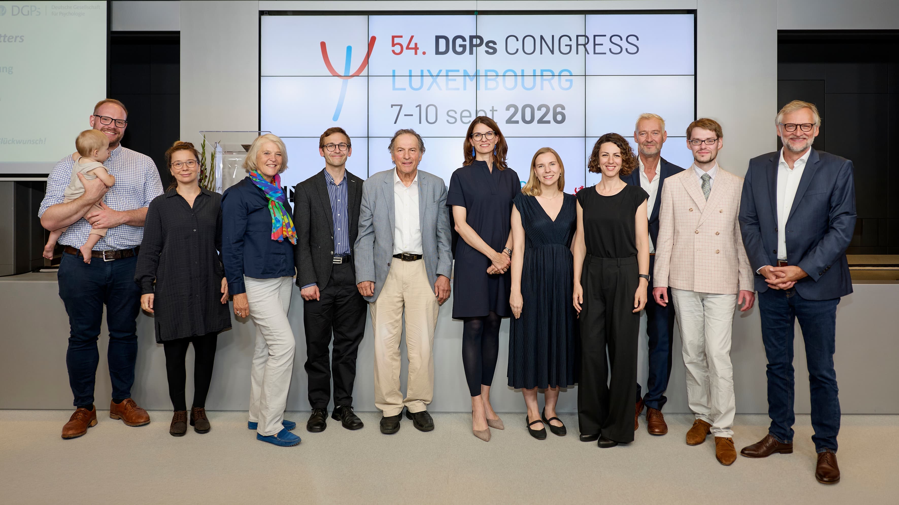 Organisers and representatives of the 54th DGPs Congress pose for a group photo in front of the congress screen displaying “Luxembourg, 7–10 September 2026”.