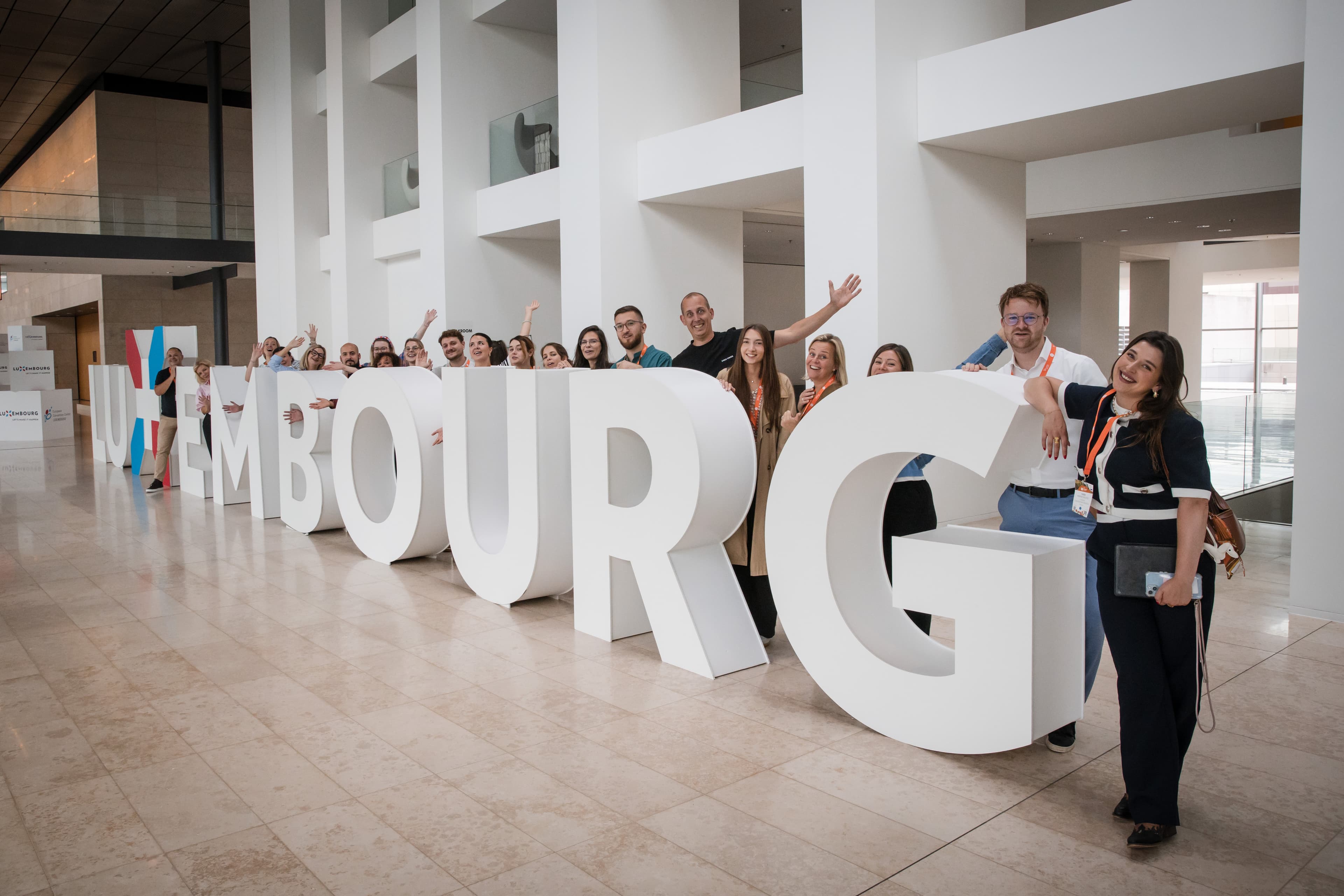 A cheerful group posing behind giant white LUXEMBOURG letters in a bright venue foyer
