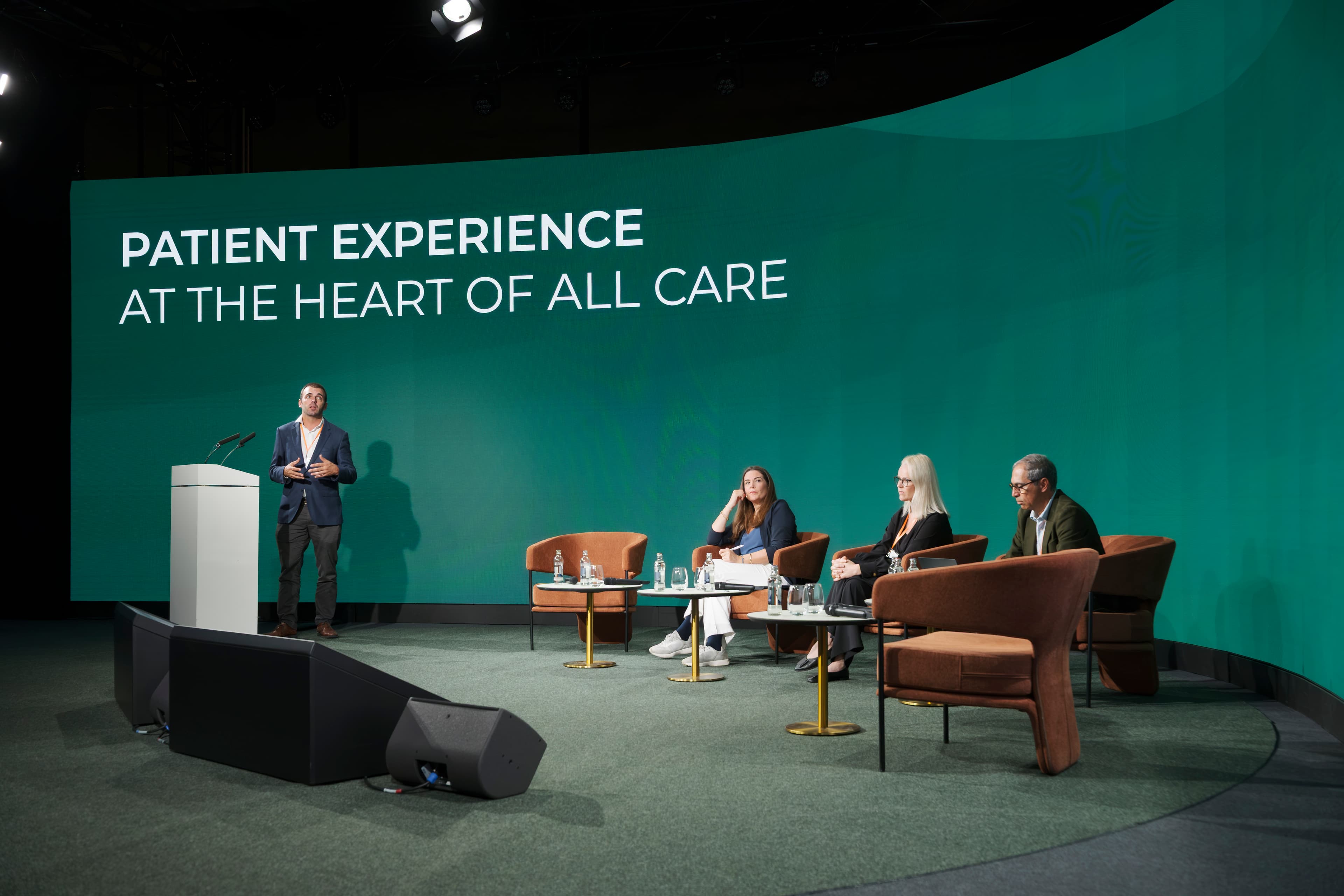 Speaker at a lectern and three seated panelists on stage under a Patient Experience screen