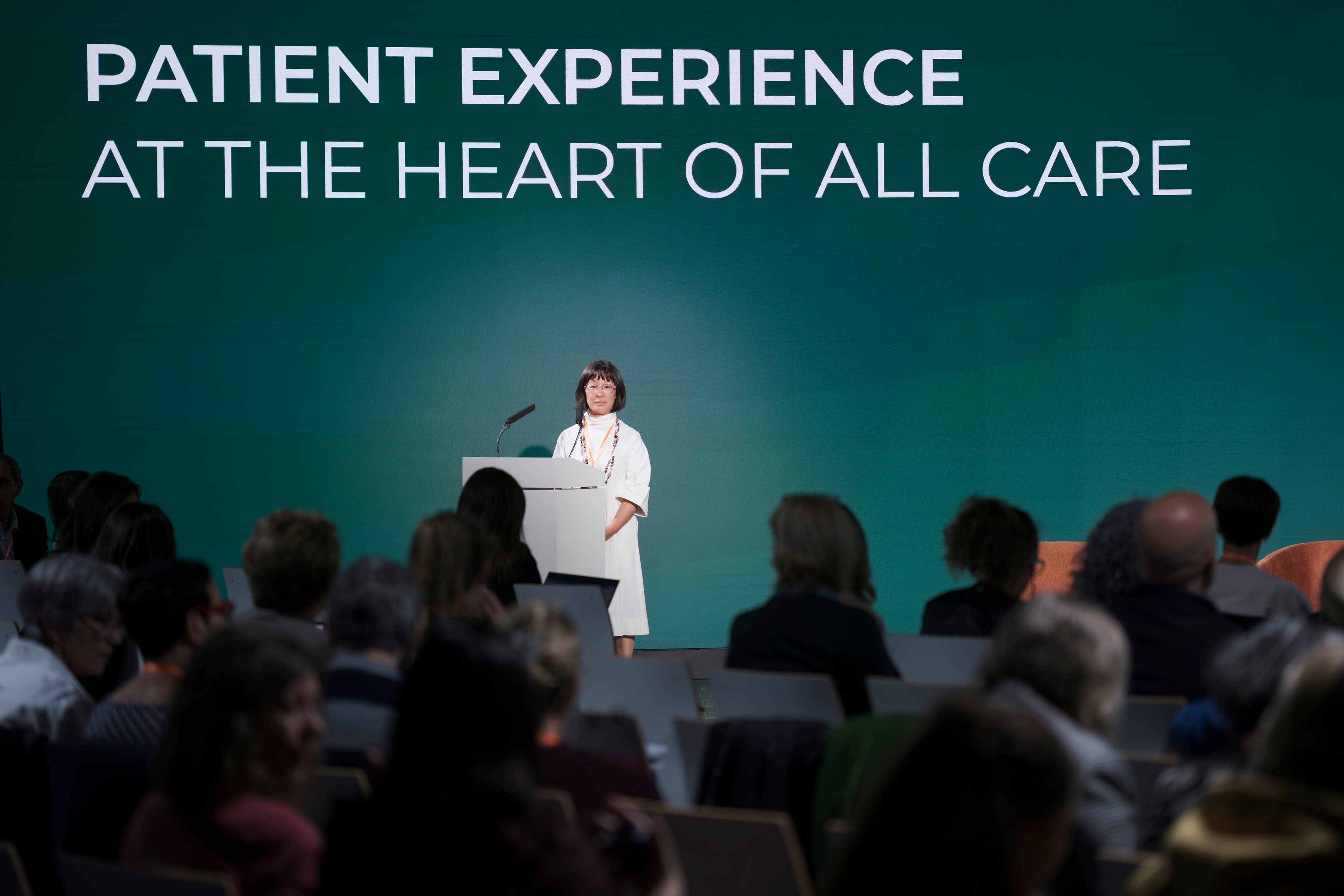 A speaker at a lectern before the audience under a Patient Experience screen at the SPX Colloquium